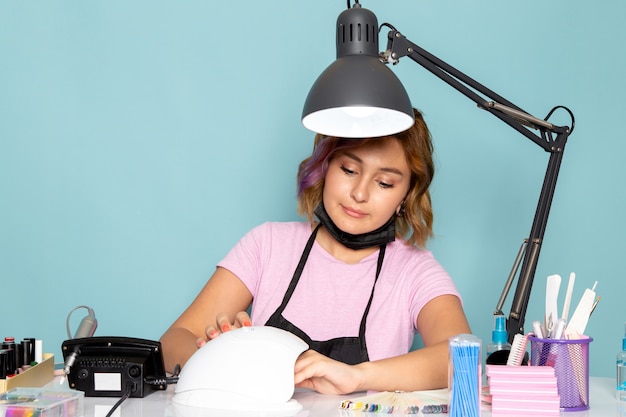 Eine junge weibliche Maniküre der Vorderansicht im rosa T-Shirt mit schwarzen Handschuhen, die vor dem Tisch sitzen und mit ihren Nägeln auf Blau arbeiten