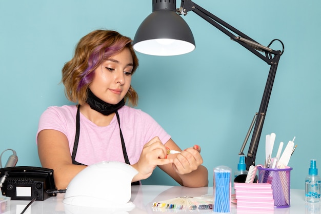 Eine junge weibliche Maniküre der Vorderansicht im rosa T-Shirt mit schwarzen Handschuhen, die vor dem Tisch sitzen und mit ihren Nägeln auf Blau arbeiten