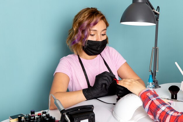 Eine junge weibliche Maniküre der Vorderansicht im rosa T-Shirt mit schwarzen Handschuhen und schwarzer Maske, die Maniküre auf Blau tut