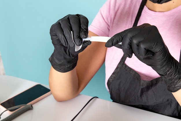 Eine junge weibliche Maniküre der Vorderansicht im rosa T-Shirt und im schwarzen Umhang mit schwarzen Handschuhen, die Maniküredetails auf Blau halten