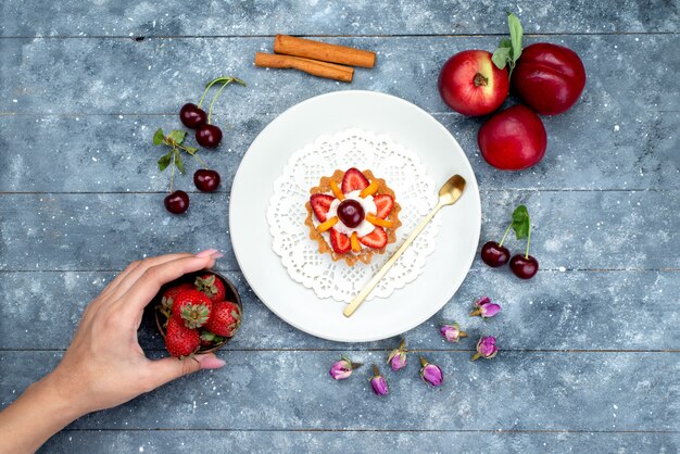 Eine kleine köstliche Torte der Draufsicht mit Sahne und Früchten innerhalb der weißen Platte zusammen mit frischen Früchten auf dem grau-blauen Hintergrundfruchtkuchen-Kekstee