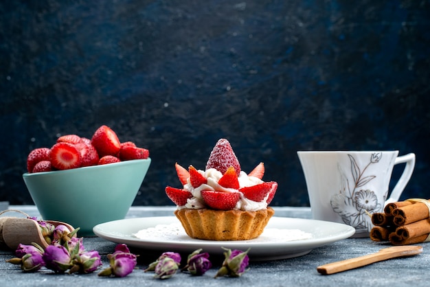 Eine kleine köstliche Torte der Vorderansicht mit Sahne-Innenplatte mit frisch geschnittenen Erdbeeren und Tee auf der blaugrauen Hintergrundplätzchenkekskuchenfrucht