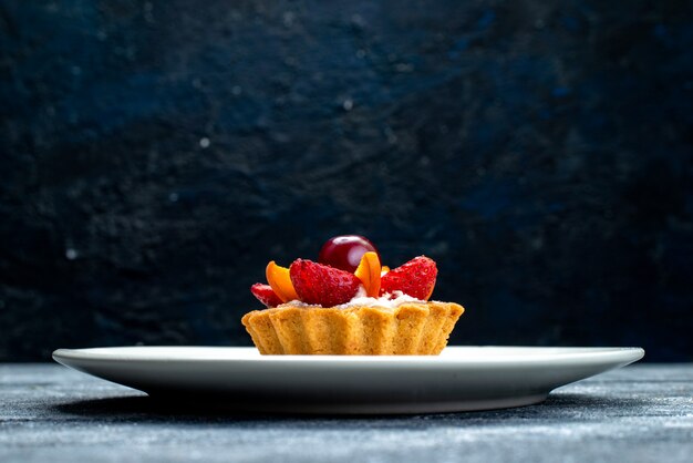 Eine kleine köstliche Torte der Vorderansicht mit Sahne und Früchten innerhalb des weißen Tellers auf dem graublauen Schreibtischfruchtkuchenplätzchen