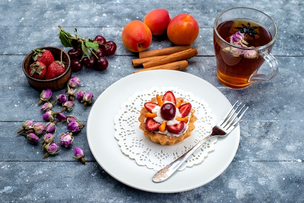 Eine köstliche kleine Torte von oben mit Sahne und frisch geschnittenen Früchten zusammen mit Zimttee auf dem grau-blauen Schreibtischfruchtkuchen-Keks