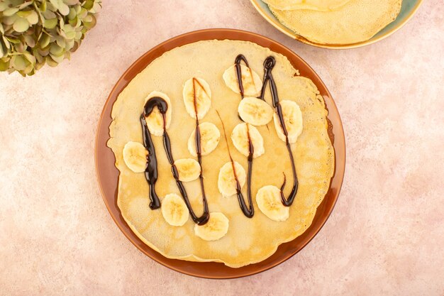 Eine köstliche Pfannkuchen der Draufsicht, die mit Schokolade und geschnittenen Bananen innerhalb des runden Tellers auf dem rosa Schreibtischnahrungsmittel-Nachtischgebäck entworfen wurde