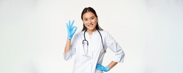 Kostenloses Foto eine lächelnde asiatische ärztin in gummihandschuhen und medizinischer uniform zeigt ein okay-zeichen und genehmigt a