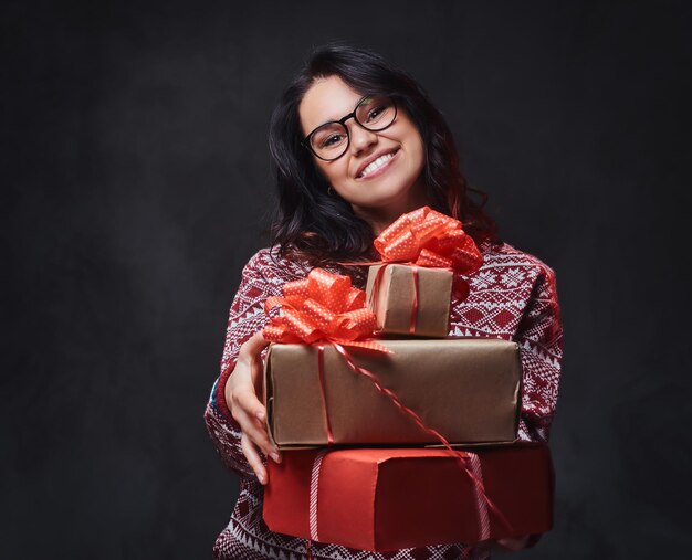 Eine lächelnde brünette Frau in rotem Pullover und Brille hält Weihnachtsgeschenke auf grauem Hintergrund.