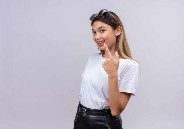 Eine lächelnde hübsche junge Frau im weißen T-Shirt, das Sonnenbrille auf ihrem Kopf trägt, zeigt mit Zeigefinger auf etwas auf einer weißen Wand