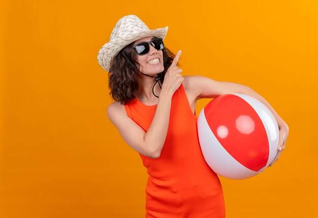 Eine lächelnde junge Frau mit kurzen Haaren in einem orangefarbenen Hemd, das Sonnenhut und Sonnenbrille hält, die aufblasbaren Ball hält, der mit Zeigefinger oben zeigt