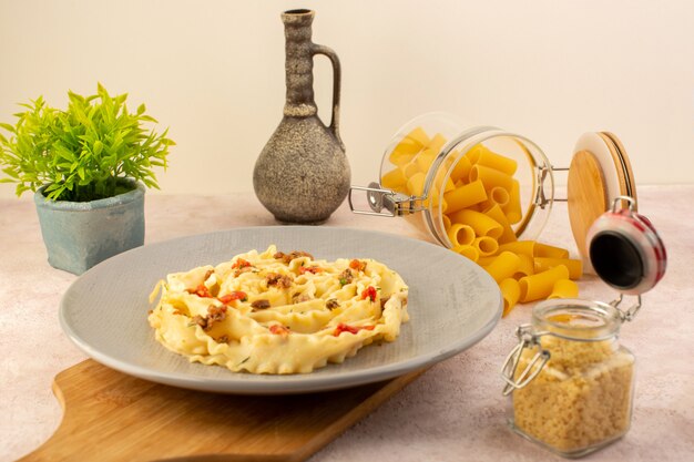 Eine leckere italienische Pasta-Mahlzeit der Vorderansicht mit gekochtem Gemüse und kleinen Fleischscheiben innerhalb der grauen Platte zusammen mit Blume und rohen Nudeln auf Rosa