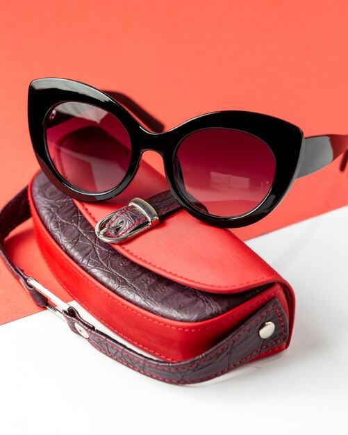 Eine moderne dunkle Sonnenbrille von vorne mit roter Ledertasche auf dem Weiß-Rot