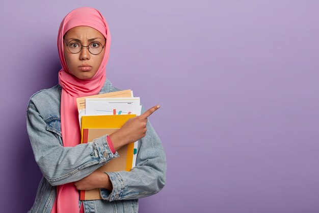 Eine muslimische Studentin im Teenageralter posiert mit Papieren und Lehrbüchern, zeigt auf freien Platz, trägt eine runde optische Brille und einen rosa Hijab