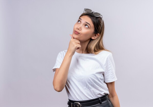 Eine nachdenkliche hübsche junge Frau im weißen T-Shirt, die Sonnenbrille auf ihrem Kopf trägt und Hand auf Kinn hält, während sie aufschaut