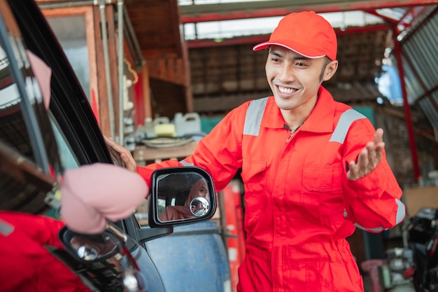 Eine nahaufnahme eines asiatischen mannes in roter uniform begrüßt mit einer willkommenen geste, als ein kunde mit dem auto zur reparatur in die werkstatt kommt