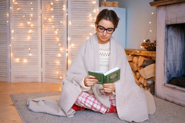 Eine nette junge Frau mit Brille sitzt am Kamin in einer Decke und liest ein Buch