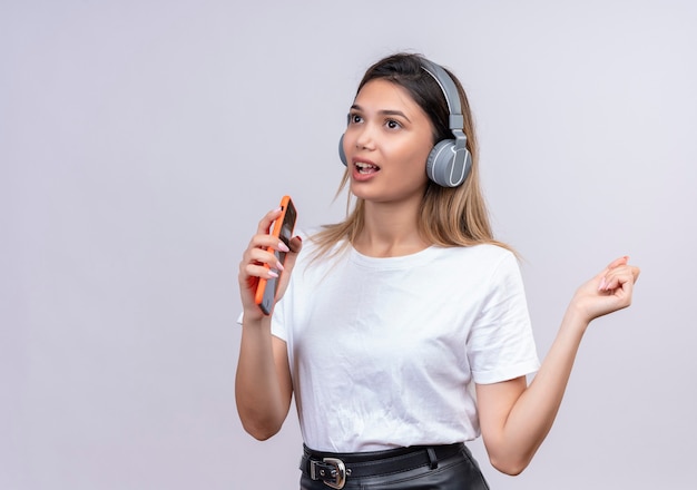 Eine niedliche junge Frau im weißen T-Shirt, das Kopfhörer trägt singend, während die Musik auf ihrem Telefon an einer weißen Wand hört