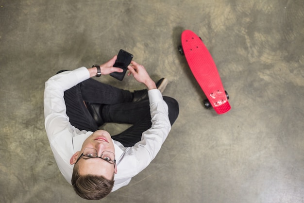 Eine obenliegende Ansicht eines jungen Mannes, der in der Hand beweglich schaut, sitzend nahe dem roten Skateboard