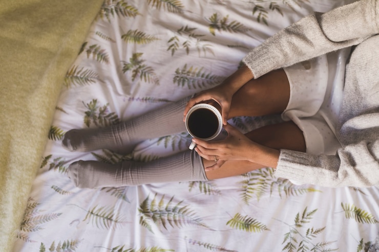 Kostenlose Foto eine obenliegende ansicht eines mädchens, das auf den tragenden socken des betts sitzt, die kaffeetasse halten