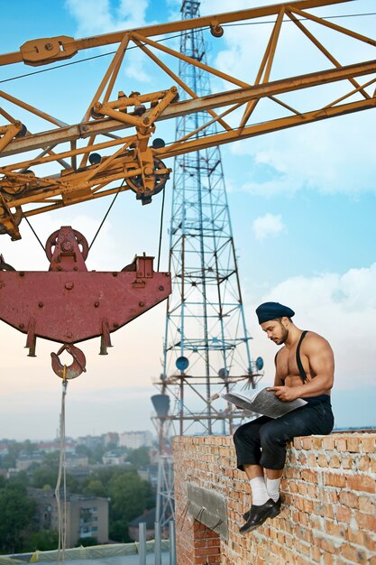 Eine Pause machen. Vertikale Aufnahme eines muskulösen Baumeisters, der eine Zeitung liest, die auf der Backsteinmauer am Bau sitzt