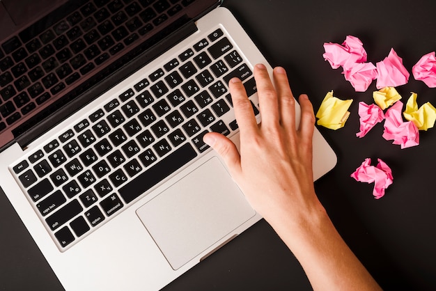 Kostenloses Foto eine person, die knopf auf laptop mit den rosa und gelben zerknitterten papieren auf schwarzem hintergrund bedrängt