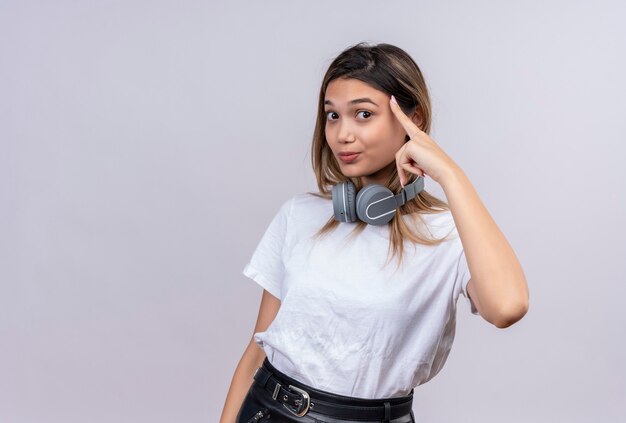 Eine positive hübsche junge Frau im weißen T-Shirt in den Kopfhörern, die auf ihren Kopf zeigen, während sie auf eine weiße Wand schauen