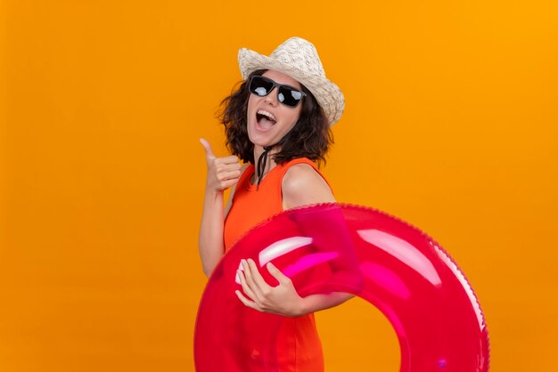 Eine positive junge Frau mit kurzen Haaren in einem orangefarbenen Hemd, das Sonnenhut und Sonnenbrille hält, die aufblasbaren Ring hält, der Daumen hoch zeigt