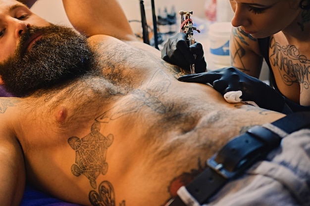 Eine professionelle Tätowiererin macht in einem Salon ein Schwarz-Weiß-Tattoo auf einem bärtigen männlichen Torso.