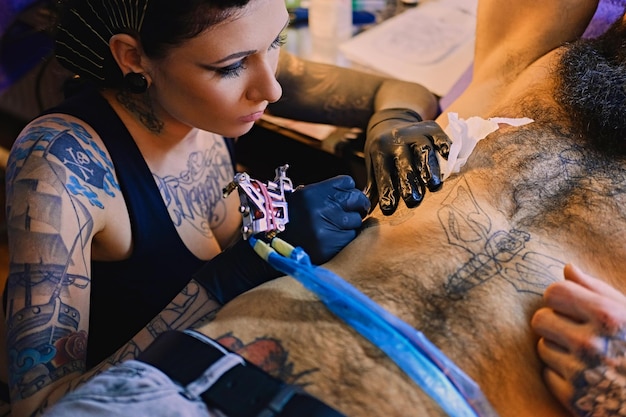 Eine professionelle Tätowiererin macht in einem Salon ein Schwarz-Weiß-Tattoo auf einem bärtigen männlichen Torso.
