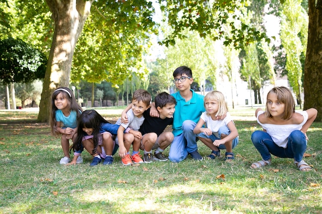 Eine Reihe glücklicher Kinder, die gemeinsam im Park Kniebeugen machen, sich umarmen und aufgeregt wegschauen. Kinderparty oder Unterhaltungskonzept