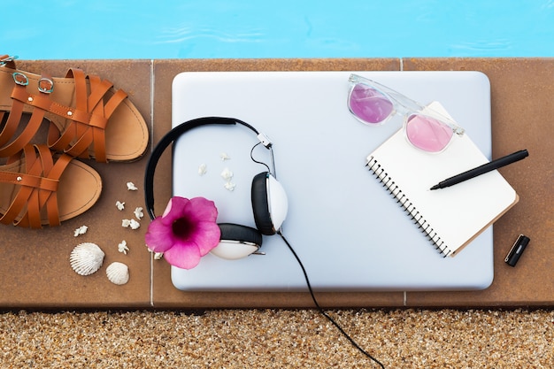 Kostenloses Foto eine reihe von hipster-freiberufler-reiseobjekten, die am pool liegen, frauensommerart, ein laptop, kopfhörer, sandalen, reisetagebuchbuch, stift, sonnenbrille, ansicht von oben, flatlay