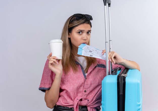 Eine reizende junge Frau, die rotes Hemd und Sonnenbrille trägt, während sie Flugtickets mit blauem Koffer auf einer weißen Wand hält