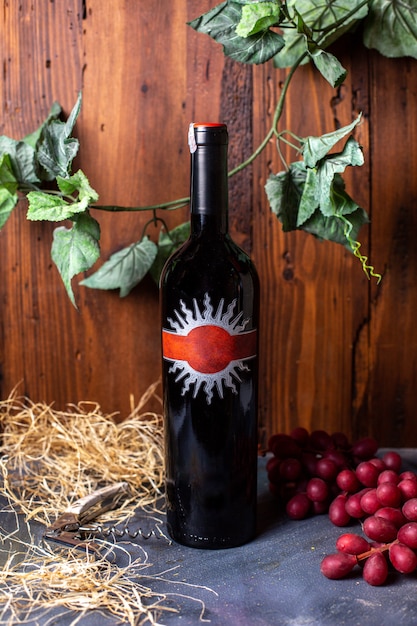 Eine Rotweinflasche Rotwein der Vorderansicht zusammen mit den roten Beeren und den grünen Blättern, die auf dem grauen Schreibtischalkohol-Weingutgetränk isoliert werden