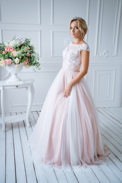 Eine schöne Braut mit Haaren und Make-up steht in einem zarten rosa Hochzeitskleid in einem hellen Dekor mit Blumen