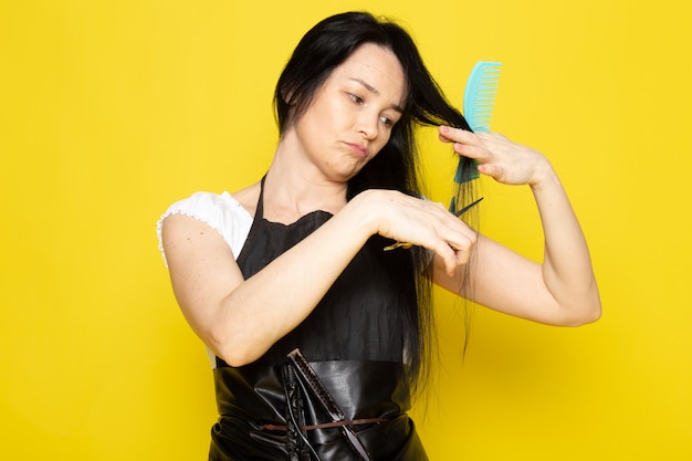 Eine schöne Friseurin der Vorderansicht im schwarzen Umhang des weißen T-Shirts mit Bürsten mit gewaschenem Haar bürstet und schneidet ihr Haar, das auf dem Friseurhaar des gelben Hintergrundstylisten aufwirft