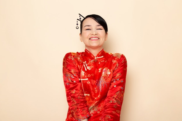 Eine schöne japanische Geisha der Vorderansicht im traditionellen roten japanischen Kleid mit den Haarstöcken, die lächelnd glücklich auf der Cremehintergrundzeremonie Japan aufwerfen