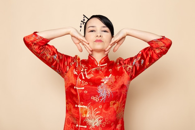 Kostenloses Foto eine schöne japanische geisha der vorderansicht im traditionellen roten japanischen kleid mit den haarstöcken, die mit ihren händen elegant auf der cremefarbenen hintergrundzeremonie darstellen, die japan ost unterhält