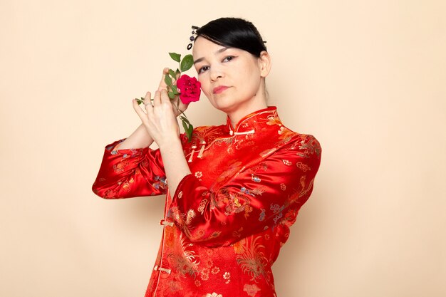 Eine schöne japanische Geisha der Vorderansicht im traditionellen roten japanischen Kleid mit Haarstäbchen, die mit roter Rose auf der cremefarbenen Hintergrundzeremonie darstellen, die Japan Ost unterhält