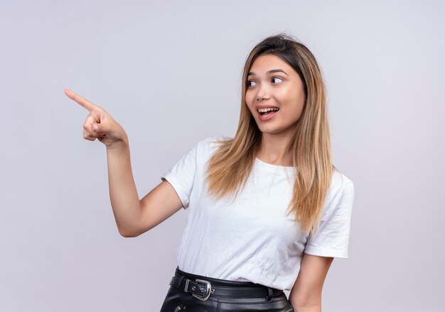 Eine schöne junge Frau im weißen T-Shirt, das mit Zeigefinger zeigt, während Seite auf einer weißen Wand schaut