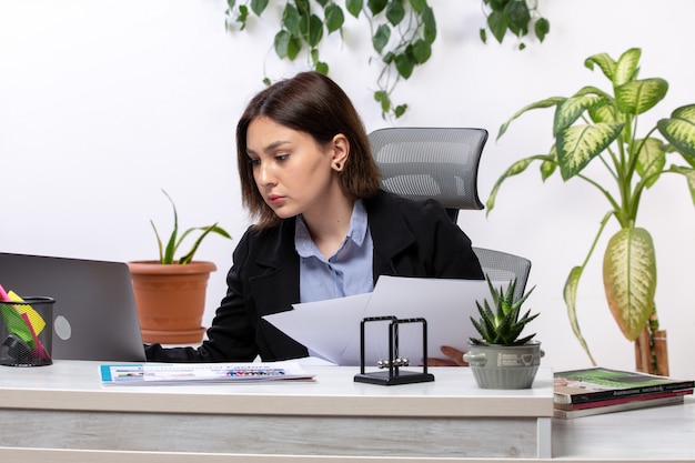 Eine schöne junge Geschäftsfrau der Vorderansicht in der schwarzen Jacke und im blauen Hemd, die mit Laptop und Dokumenten vor dem Jobbüro des Tischgeschäfts arbeiten