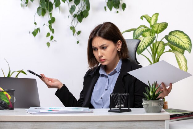 Eine schöne junge Geschäftsfrau der Vorderansicht in der schwarzen Jacke und im blauen Hemd, die mit Laptop vor dem Jobbüro des Tischgeschäfts arbeiten