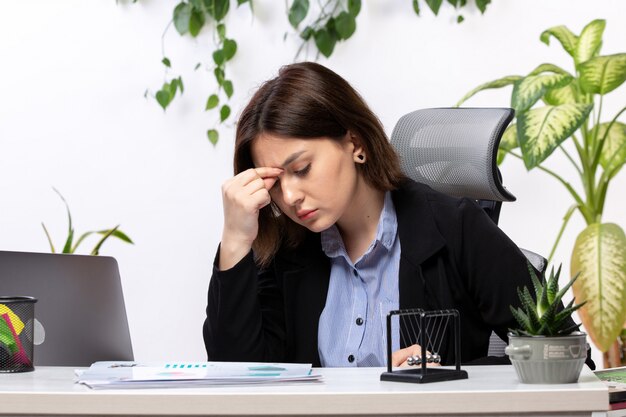 Eine schöne junge Geschäftsfrau der Vorderansicht in der schwarzen Jacke und im blauen Hemd, die sich vor dem Jobbüro des Tischgeschäfts krank fühlen