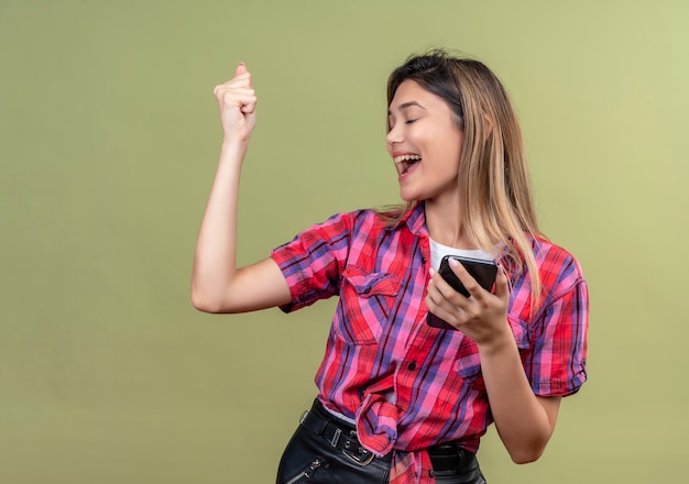 Eine sehr glückliche schöne junge Frau in einem karierten Hemd, das Handy hält, während geballte Faust erhebt