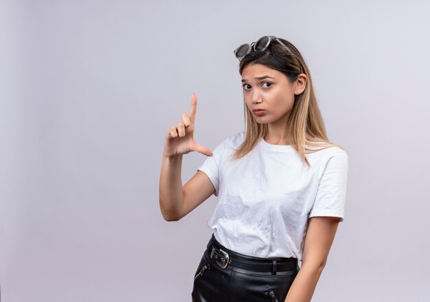 Eine selbstbewusste hübsche junge Frau im weißen T-Shirt, das Sonnenbrille auf ihrem Kopf trägt und auf eine weiße Wand schaut