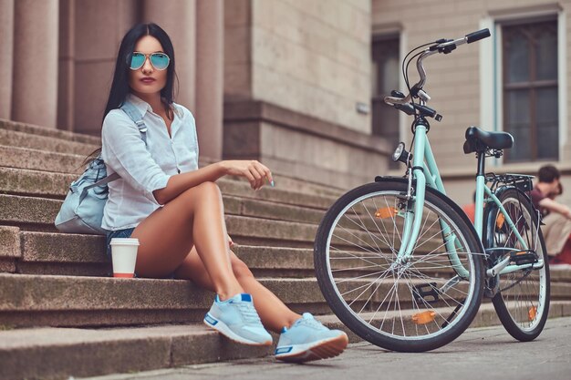 Eine sexy brünette Frau mit Bluse und Jeansshorts in Sonnenbrille, die sich nach einer Fahrradtour entspannt und mit einer Tasse Kaffee auf Stufen in einer Stadt sitzt.