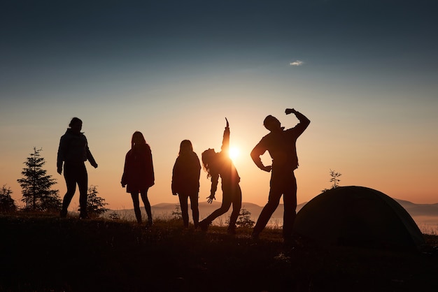 Eine Silhouette von Gruppenleuten hat Spaß auf der Spitze des Berges in der Nähe des Zeltes während des Sonnenuntergangs.