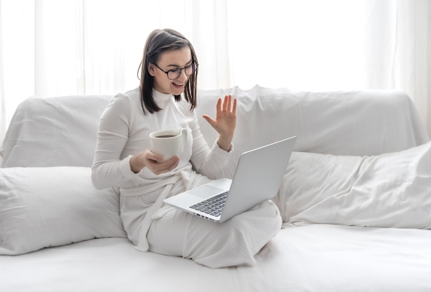 Eine süße junge Frau sitzt zu Hause auf einem weißen Sofa in einem weißen Kleid vor einem Laptop. Fernarbeit und freiberufliche Tätigkeit.