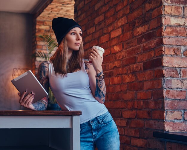 Eine tätowierte sexy Blondine in einem T-Shirt und einem Hut mit einer Tasse Kaffee und einem Tablet-Computer in einem Raum mit Loft-Interieur.