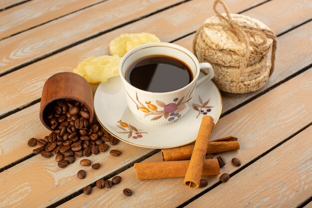 Eine Tasse Kaffee von vorne, heiß und stark mit frischen braunen Kaffeesamen, Zimt und Crackern auf dem cremefarbenen rustikalen Schreibtischkaffee-Samengetränk-Fotokorn