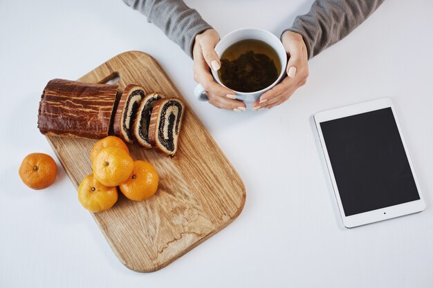 Eine Tasse Tee lässt den Morgen heller aussehen. Moderne Frau, die Topf hält, Mandarine und gerollten Kuchen während des Frühstücks oder Mittagessens isst und darauf wartet, dass ihr Chef ihre Informationen über digitales Tablet sendet