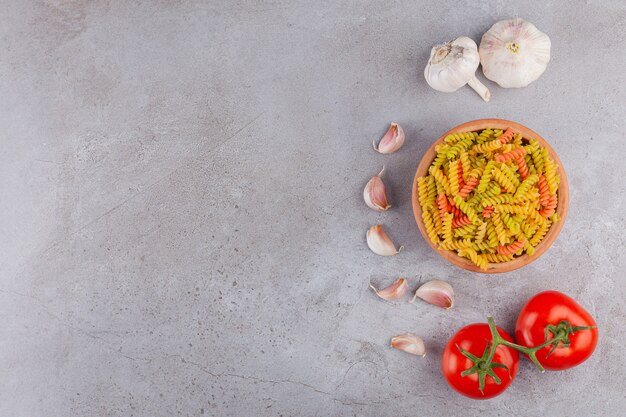 Eine Tonschale mit mehrfarbigen rohen Spiralnudeln mit Knoblauch und frischen roten Tomaten.